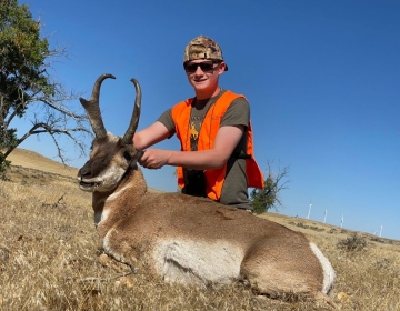 A young hunter wearing an orange safety vest and sunglasses proudly displays a pronghorn antelope harvested during an SNS Outfitter & Guides expedition.